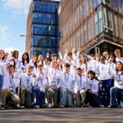 Group of secondary school students wearing white tops and lanyards pose together outside Stripe&rsquo;s Dublin office during the Young Scientist Business Bootcamp, with modern glass buildings in the background.