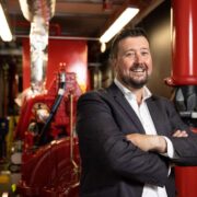 Man in suit standing with arms crossed inside a fire protection plant room, wearing a dark suit and white shirt, with red fire suppression pipes and equipment visible in the background.