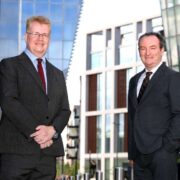 Two people wearing dark business suits stand outdoors in an urban setting, positioned several feet apart, with modern glass and steel office buildings behind them.