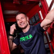Person wearing a navy TRYKA T‑shirt stands inside a red London telephone box, holding a handset to one ear and extending an arm toward the open door, with street reflections visible on the glass panels.
