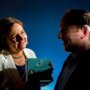 Woman holding a Vertigenius medical device package speaks with a man wearing a Vertigenius head sensor, illustrating remote monitoring technology for vertigo care.