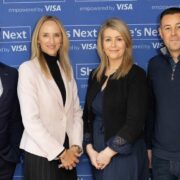 Four people standing in front of a blue backdrop with the text 'She's Next empowered by Visa.' From left to right: a smiling man in a navy suit and tie; a woman with long blonde hair wearing a light pink blazer over a black top; a woman with shoulder‑length blonde hair wearing a dark dress and cardigan; and a man in a navy half‑zip sweater and jeans.