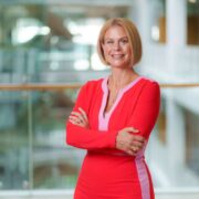 Person standing indoors with arms crossed, wearing a red outfit with pink trim, photographed in a modern office setting with glass railings and blurred background.