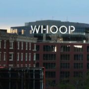 Whoop logo illuminated on the rooftop of a modern office building overlooking brick buildings in Boston at dusk.
