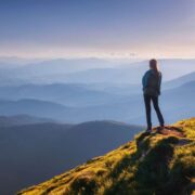 Person standing on a grassy mountain ridge overlooking layered blue hills and valleys under a clear sky, with sunlight illuminating the landscape.