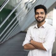 Young man standing outdoors with arms crossed in front of a modern glass building, with two people walking in the background.