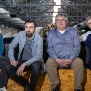 Four men seated side by side on a wooden bench in a modern office space, facing the camera.