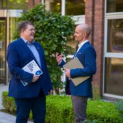 Two business professionals stand outside a modern office building, speaking and holding documents, including a copy of the Ireland Innovation Index report, with greenery and glass entrance in the background.