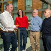 Four adults standing in a modern office atrium with glass panels and wooden walls, talking together beside a balcony railing. One person in a red sweater stands between three others dressed in neutral-toned shirts and trousers.