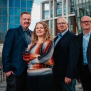 Four adults standing outdoors in front of modern glass office buildings, wearing business attire, with one person in a colourful striped sweater and three others in dark suits, posing together in an urban business district setting.