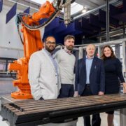 Four people standing in a modern industrial lab beside a large orange robotic arm and a metal workstation, with glass enclosures and manufacturing equipment in the background.