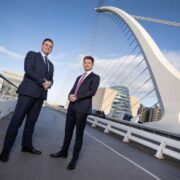Two business professionals standing on the Samuel Beckett Bridge in Dublin, with the city skyline and the bridge&rsquo;s white harp-shaped structure in the background.