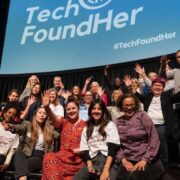 Group photograph of attendees on a stage beneath a large screen displaying the TechFoundHer logo and hashtag. Several people are seated and standing, many with raised hands, at a TechFoundHer event in a theatre-style venue.