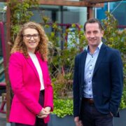 Two business professionals standing outdoors in a landscaped office courtyard, wearing smart business attire, with modern office buildings and greenery in the background.