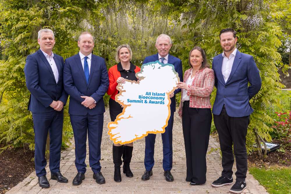 Group of six individuals standing outdoors beneath a leafy archway, holding a sign shaped like the island of Ireland displaying the text All Island Bioeconomy Summit & Awards, with greenery, flowers, and a garden path in the background.