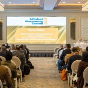 Audience seated in a conference room facing a large screen displaying the All-Island Bioeconomy Summit 2025, with a speaker presenting from a podium in a formal event setting.