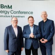 Three men in business attire stand in front of a branded backdrop reading BnM Energy Conference Infrastructure and Investment, posing for a photo at an industry event held at a conference venue.