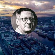 Aerial view of Dublin Docklands at sunset with the River Liffey, port and city skyline in the background, overlaid with a circular black-and-white headshot of a bespectacled person centred on the image.