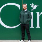 Person standing outside a green industrial unit, wearing a dark green Cl&iacute;r jacket, positioned in front of large white Cl&iacute;r branding and a bird logo on the wall.