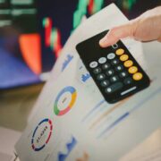Business professional holding a smartphone calculator over printed financial charts and reports with pie charts and graphs, symbolising accounting, credit control, or debt analysis in an office setting.