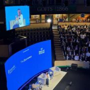 Wide shot of a conference auditorium with tiered seating filled with attendees watching a live interview on stage. A speaker sits in an armchair on a lit platform beside a small table, while a large blue screen behind displays “The Currency” and “Bank of Ireland.” An overhead screen shows a close-up of the on-stage conversation. The venue signage “GOFFS 1866” and “The Real Deal” is visible in the background.