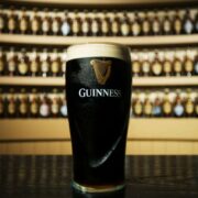 A pint of Guinness with a creamy head stands on a bar counter, with rows of bottled Guinness displayed on illuminated shelves in the background.