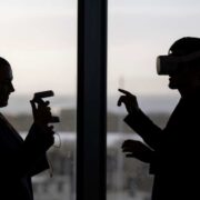 Silhouetted view of two people indoors by a window, one wearing a virtual reality headset and gesturing, the other holding a handheld device, with a cityscape visible in the background.