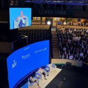 A large conference audience seated in a tiered indoor venue watches a speaker seated on stage. A curved digital screen displays event branding and sponsor logos, while a live video feed of the speaker appears on a screen above the seating area.