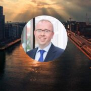 Aerial view of Dublin Docklands at sunset with modern office buildings and waterfront, overlaid with a circular headshot of a professional in business attire at the center of the image.