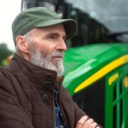 Farmer standing beside a green tractor in a rural setting, wearing a cap and jacket with arms folded, looking across farmland.