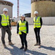Three people wearing high-visibility vests and hard hats stand on a gravel construction site in front of large cylindrical industrial tanks, with machinery and a crane visible in the background under a clear sky.