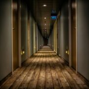 Narrow, softly lit hotel corridor with wooden flooring, lined with identical closed guest room doors on both sides and overhead lighting leading to a vanishing point at the far end.