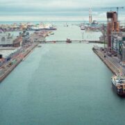 Aerial view of the River Liffey flowing through Dublin Docklands, with modern office buildings, construction cranes, bridges, quayside roads and a cargo vessel under an overcast sky.