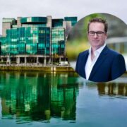Portrait-style composite image showing a modern glass-fronted office building beside a waterfront, with reflective green-blue water in the foreground, and an overlaid circular headshot of a suited business professional positioned to the right.