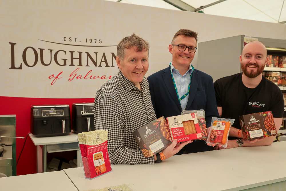 Three individuals stand behind a Loughnane’s of Galway exhibition counter at Bord Bia Bloom, holding packaged meat products including sausages and prepared dishes, with branded signage, food items on display and kitchen equipment visible in the background.