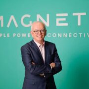 Man in a navy suit standing with arms crossed in front of a green backdrop displaying the “MAGNET+” logo and the phrase “People Powered Connectivity”.