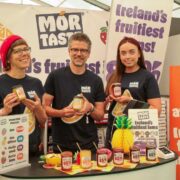 Three people stand at a Mór Taste display booth at Bord Bia Bloom, holding jars of fruit spreads and jams, with branded signage, product samples, and a price list visible in the background.