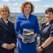 Three people standing outdoors on a rooftop or terrace overlooking a city, with hills in the background, holding a report titled &ldquo;The New Retirement Reality,&rdquo; a study on retirement preparedness in Ireland.