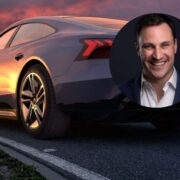 Electric car parked on a roadside at sunset, viewed from the rear side, with warm orange light reflecting off the vehicle’s surface; circular inset shows a professional headshot of a person in formal attire overlaid on the image.