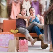 Two shoppers seated on an orange bench in a clothing store surrounded by shopping bags, pointing toward a standing shopper holding garments while racks of clothes hang in the background.
