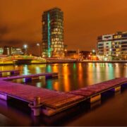 Night-time view of Dublin&rsquo;s Silicon Docks with illuminated modern buildings reflecting on calm water, a lit pedestrian bridge crossing the river, and floating wooden platforms in the foreground under an orange city sky.