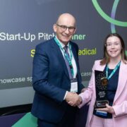 Two people stand on a stage at Enterprise Ireland&rsquo;s Start-Up Day event, shaking hands as one accepts the Spinout Showcase Award trophy, with a presentation screen visible in the background.