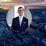 Aerial cityscape at sunset with a circular inset portrait of a business executive in a suit, set against an urban waterfront skyline.