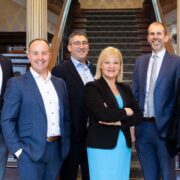 Group of six business professionals standing together in a formal interior setting with a staircase background, dressed in smart business attire, representing Xeinadin and Carrigan O’Dwyer following their partnership announcement.