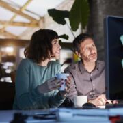 Two people seated at a desk in a modern office, collaborating in front of a large computer monitor with sticky notes on the screen, holding coffee cups, representing teamwork and AI-driven content creation.