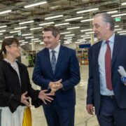 Three business professionals in formal attire walking and talking inside a large, brightly lit manufacturing facility, with industrial equipment, shelving units and production lines visible in the background, one person holding documents while gesturing during the discussion.