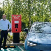 Person standing beside a red EZO electric vehicle charging unit in an outdoor car park, with a blue electric car plugged in and trees in the background.