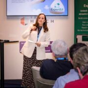 Presenter speaking at an ExpertEd launch event at The Hatch Lab in Gorey, standing beside a screen and branded banner while addressing a seated audience about the eLearning platform and its features.