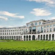 Exterior view of a large luxury hotel with a curved cream-coloured façade, tall arched windows, and landscaped gardens with a lawn and fountain under a partly cloudy sky.