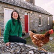 Person sitting on a low stone wall outside a traditional stone cottage with a red door and white-framed windows, wearing a green sweater, with a brown chicken walking along the wall in the foreground and a grassy garden surrounding the house.